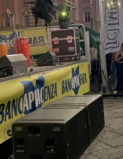 Bambina che canta sul palco durante un evento serale a Piacenza, osservata da un’altra bimba tra il pubblico