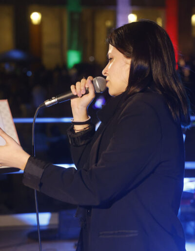 Cantante di ArteMusica Piacenza si esibisce dal vivo leggendo il testo da un leggio durante un concerto serale