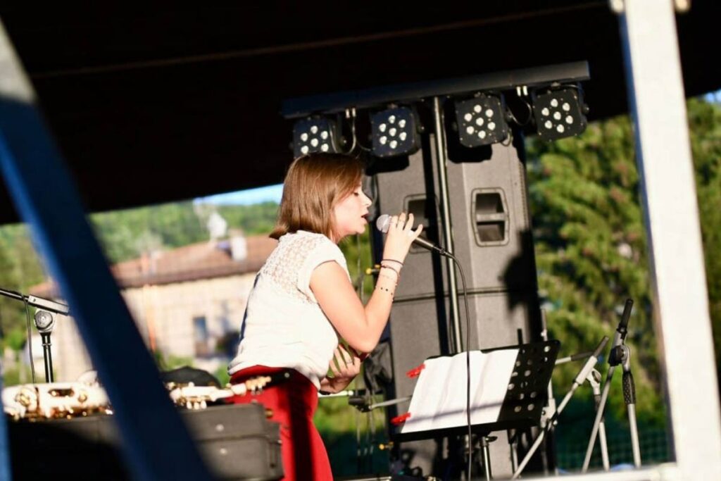 Giovane cantante si esibisce dal vivo su un palco all’aperto con microfono e leggìo