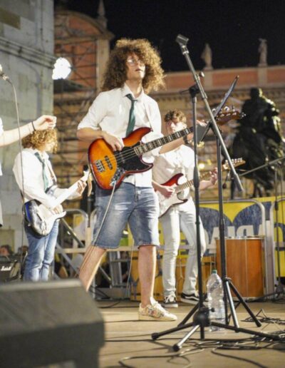 Giovane bassista suona dal vivo con una band di ragazzi durante un concerto serale in piazza a Piacenza.