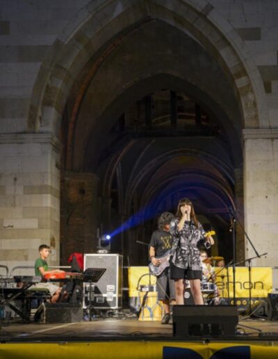 Concerto serale a Piacenza con giovani musicisti sul palco e una cantante durante l'esibizione sotto i portici del centro storico.