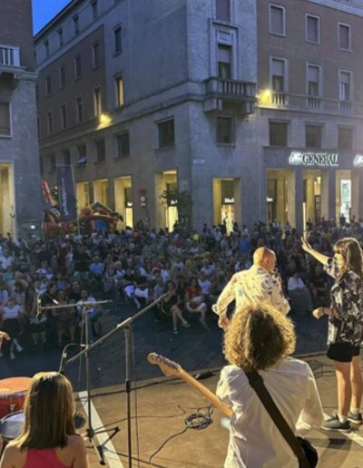 Concerto delle scuole di musica a Piacenza visto dal palco, con giovani musicisti e grande pubblico in piazza durante un evento serale.