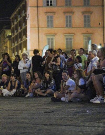 Pubblico seduto e in piedi che assiste a un evento serale nel centro storico
